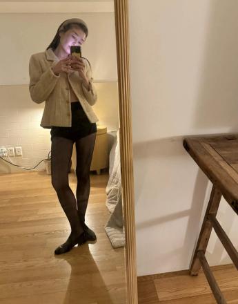A person in a tweed jacket, black shorts, and sheer tights takes a mirror selfie.