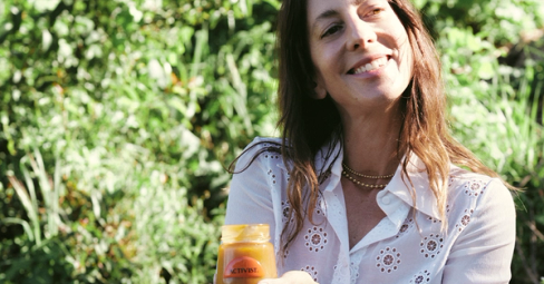 Smiling woman in a white shirt holding a jar of Activist Manuka Honey against a green background.