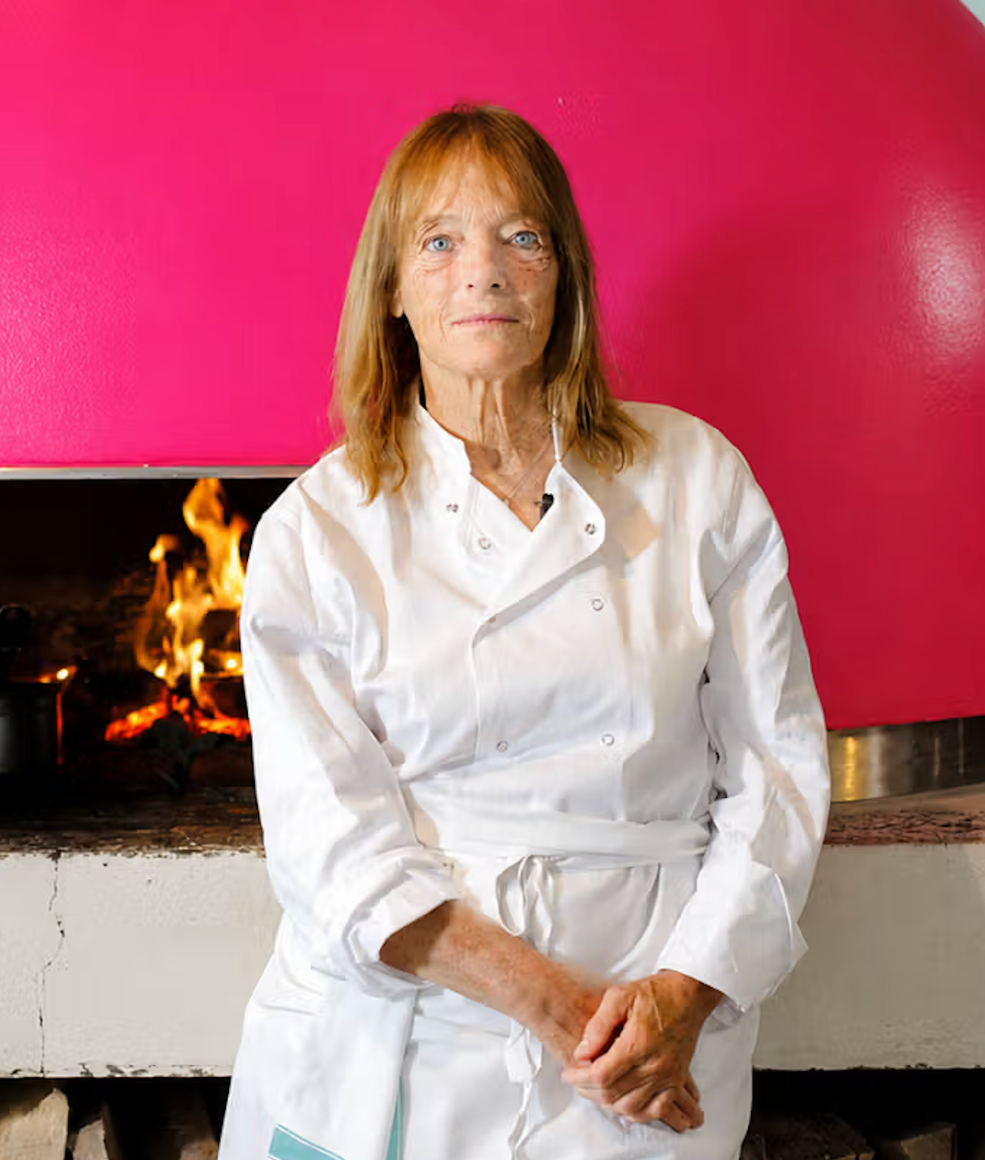 A woman in a chef's uniform stands in front of a bright pink pizza oven with fire burning inside.