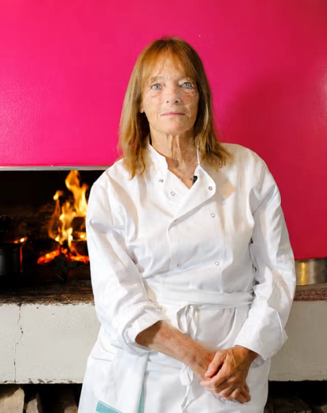 A woman in a chef's uniform stands in front of a bright pink pizza oven with fire burning inside.