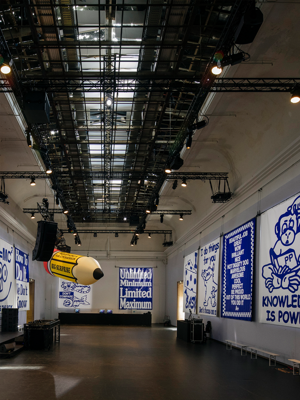 Large event hall with a grid ceiling, stage lights, blue and white graphic banners on the walls, and a giant inflatable yellow pencil in the center.