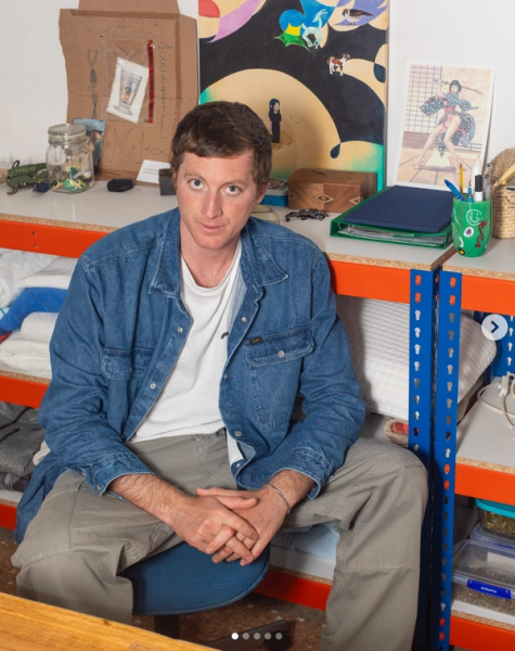 A man in a denim shirt and khaki pants sits looking at the camera, with a workbench and shelves displaying art and various items behind him.