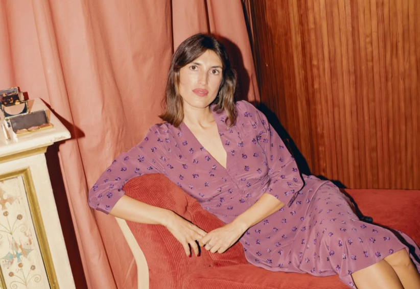 A woman in a purple floral dress reclines on a red sofa, with a pink curtain and wood paneling behind her.