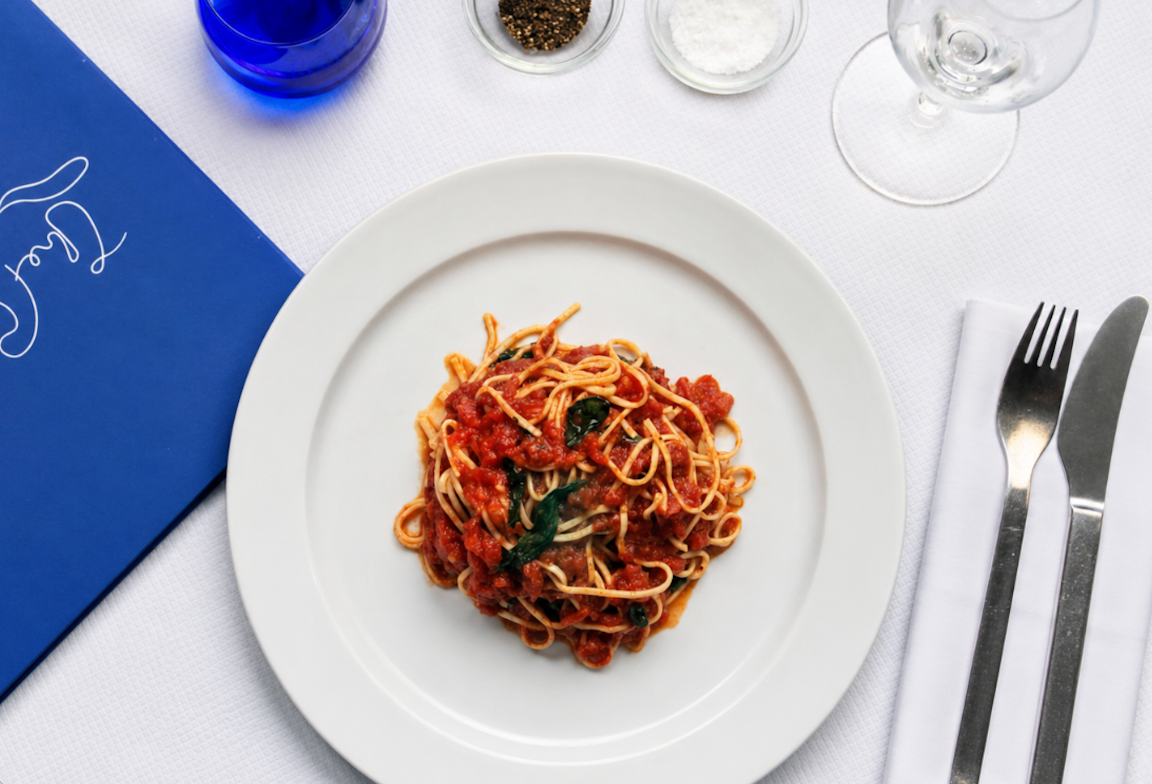 Spaghetti with tomato sauce and basil on a white plate, at a restaurant table with a blue menu, silverware, and condiments.