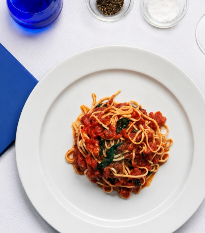 Spaghetti with tomato sauce and basil on a white plate, at a restaurant table with a blue menu, silverware, and condiments.