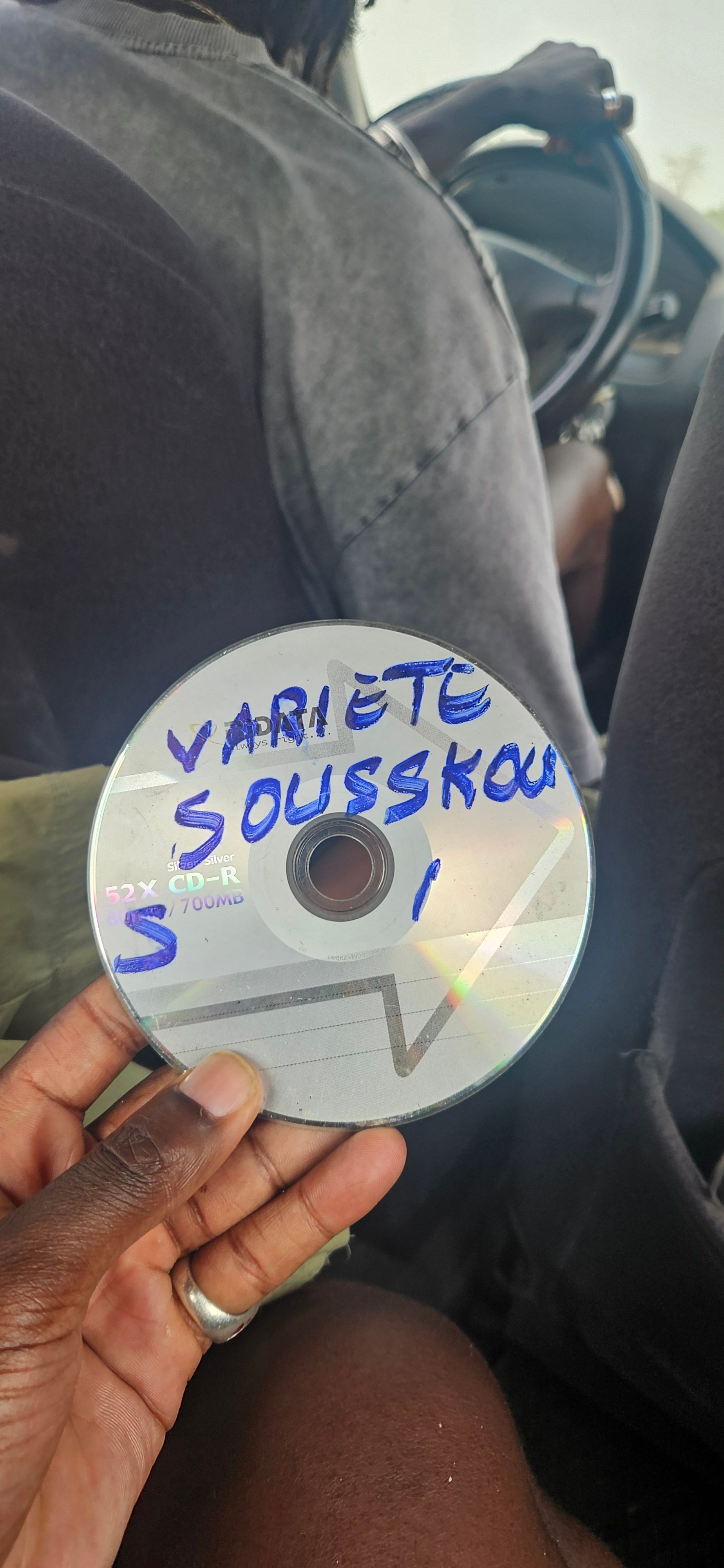 A hand holds a CD-R labeled "VARIETE SOUSSKOU S / 1" in blue marker, inside a car.