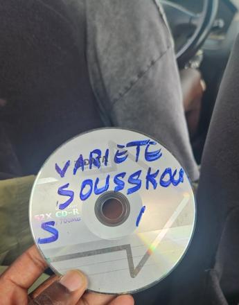 A hand holds a CD-R labeled "VARIETE SOUSSKOU S / 1" in blue marker, inside a car.
