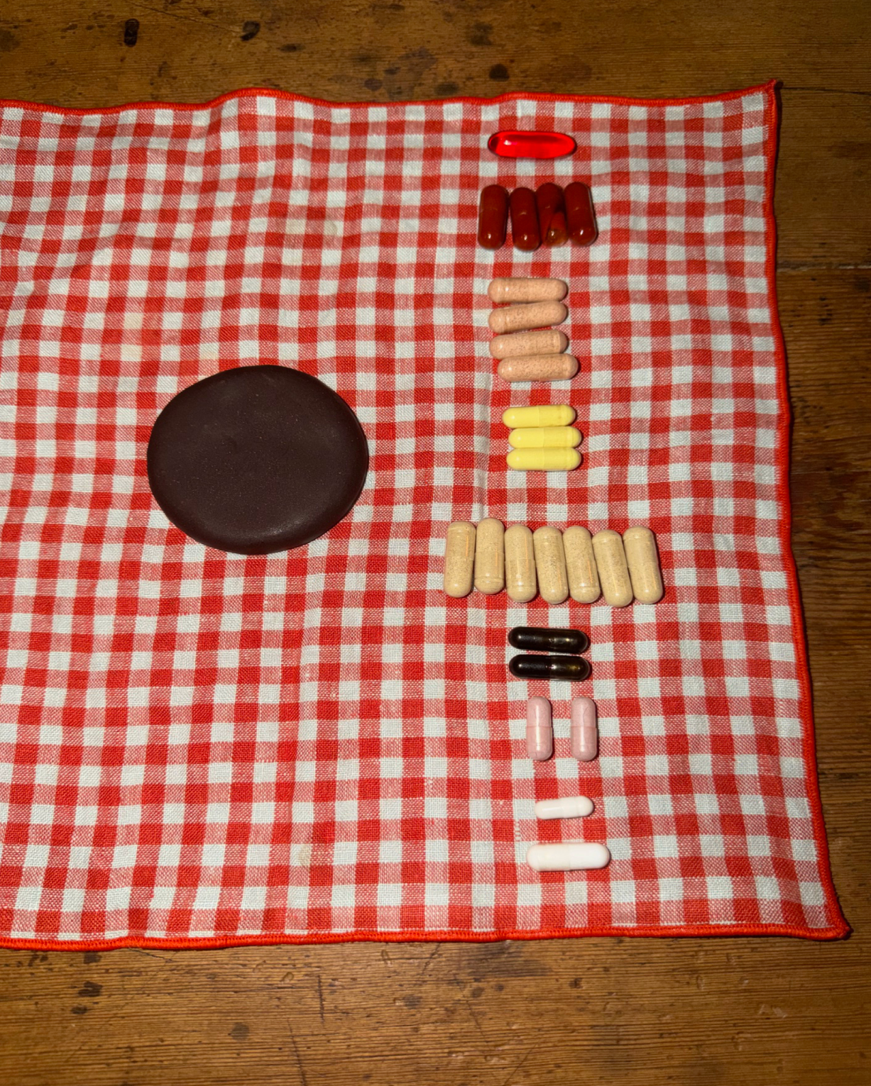 A dark chocolate disc next to several rows of colorful pills on a red and white checkered cloth.