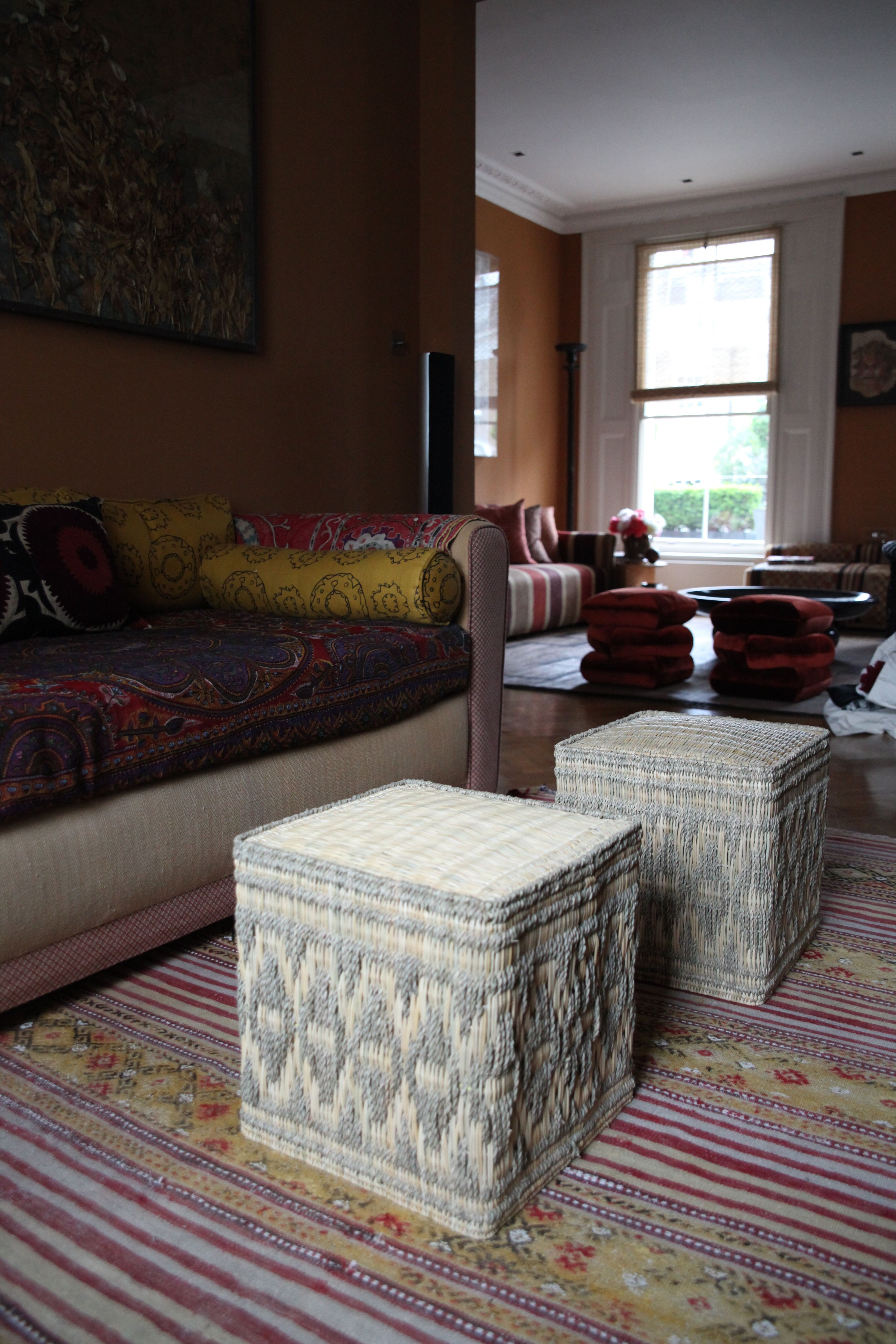 A richly decorated living room with patterned sofas, woven ottomans, and a large window.