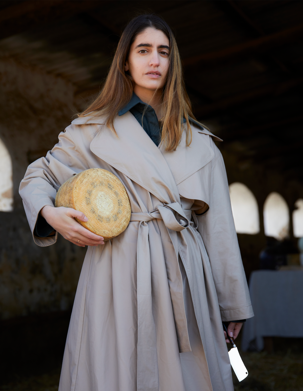 A woman in a beige trench coat holds a round cheese and a cleaver, standing in a rustic barn.