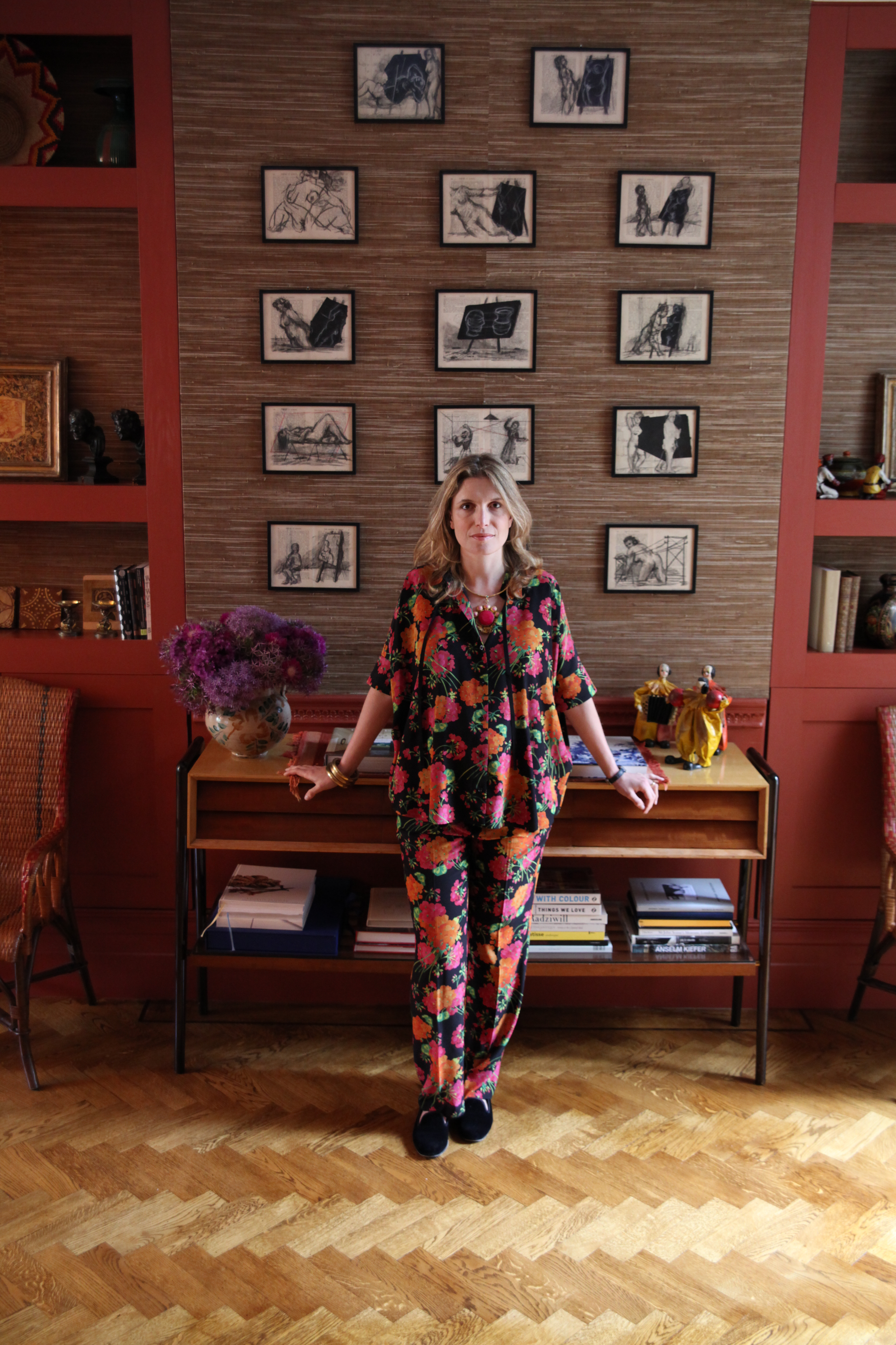 A woman in a vibrant floral outfit stands in a room with a textured wall displaying a grid of abstract art, flanked by red bookshelves.