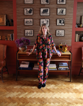 A woman in a vibrant floral outfit stands in a room with a textured wall displaying a grid of abstract art, flanked by red bookshelves.