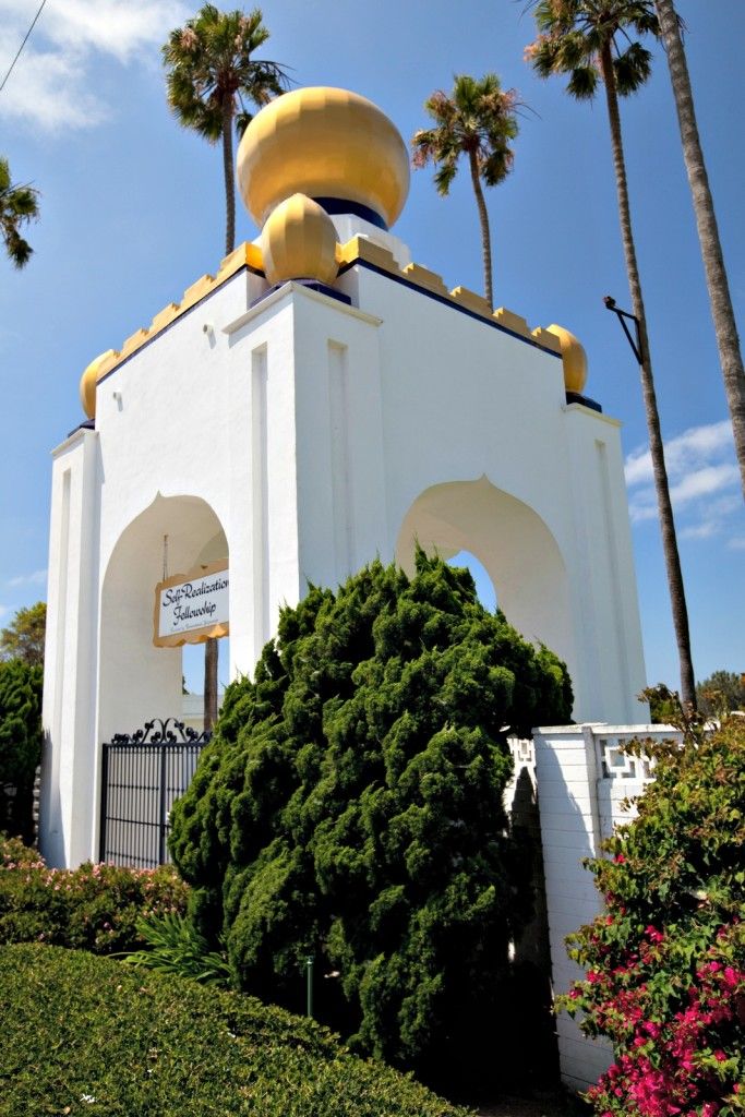 The Self-Realization Fellowship building, a white structure with golden domes and arches, surrounded by lush gardens and tall palm trees.