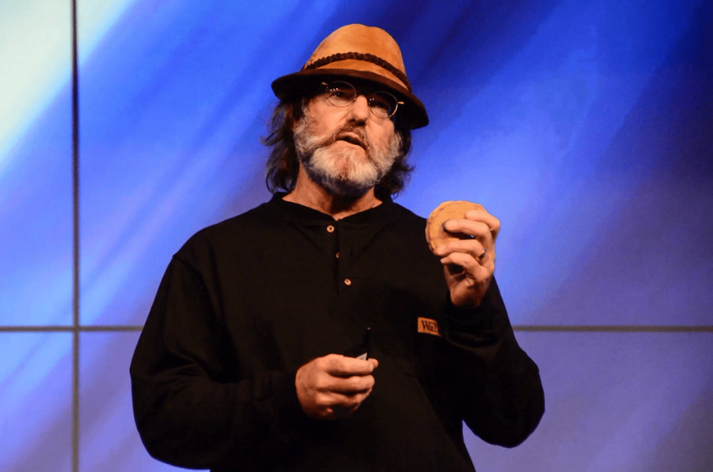 A man with a beard, glasses, and a brown hat holds a round food item and a pen, standing against a blue background.