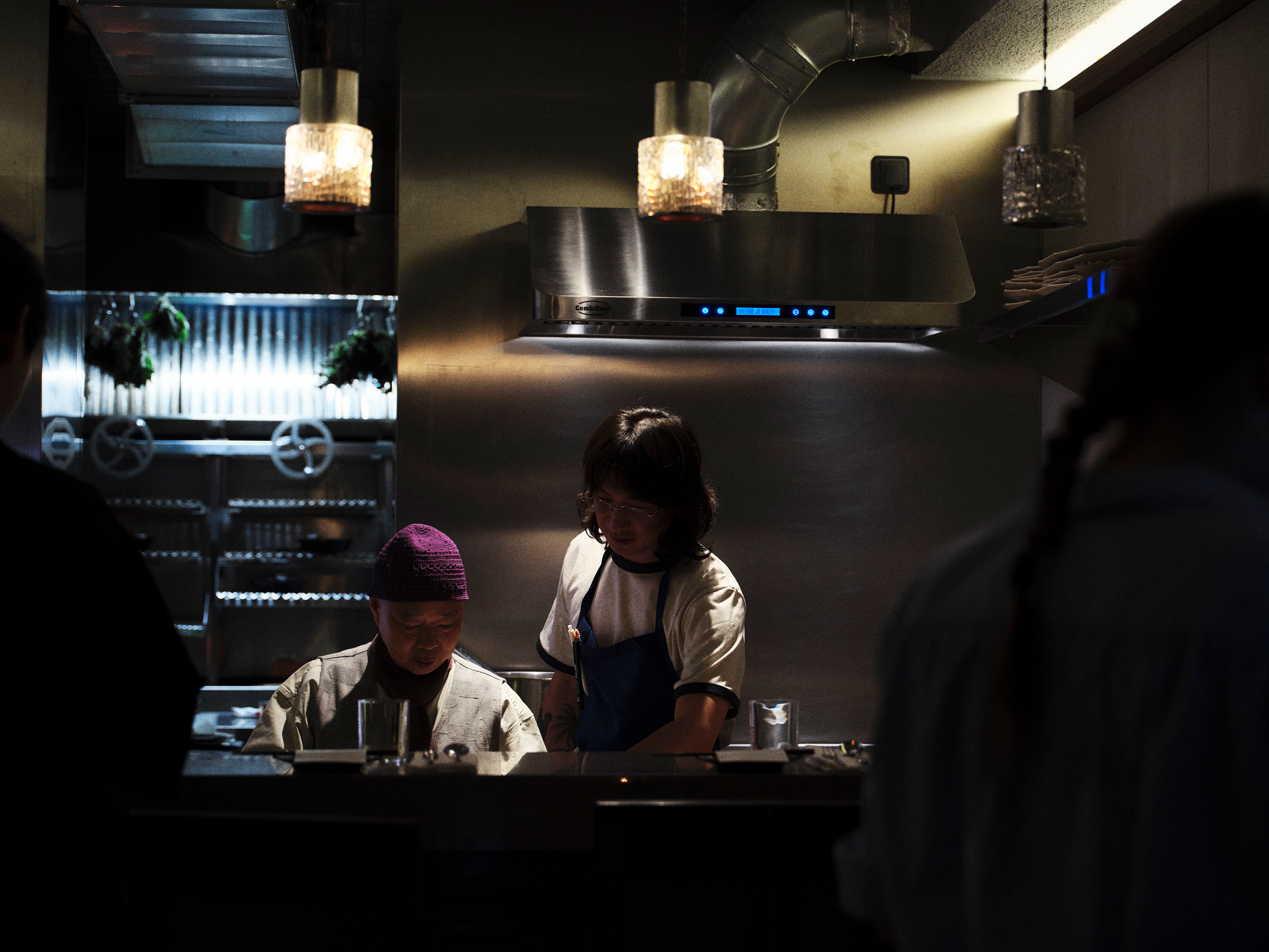 Two chefs work behind a stainless steel counter in a dimly lit restaurant kitchen.
