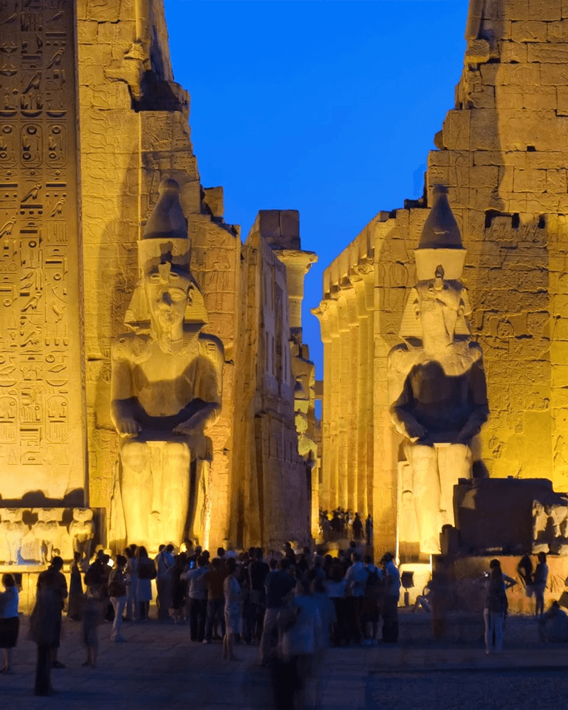 Luxor Temple at night, with two colossal pharaoh statues illuminated by golden light, and a crowd of visitors.