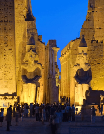 Luxor Temple at night, with two colossal pharaoh statues illuminated by golden light, and a crowd of visitors.