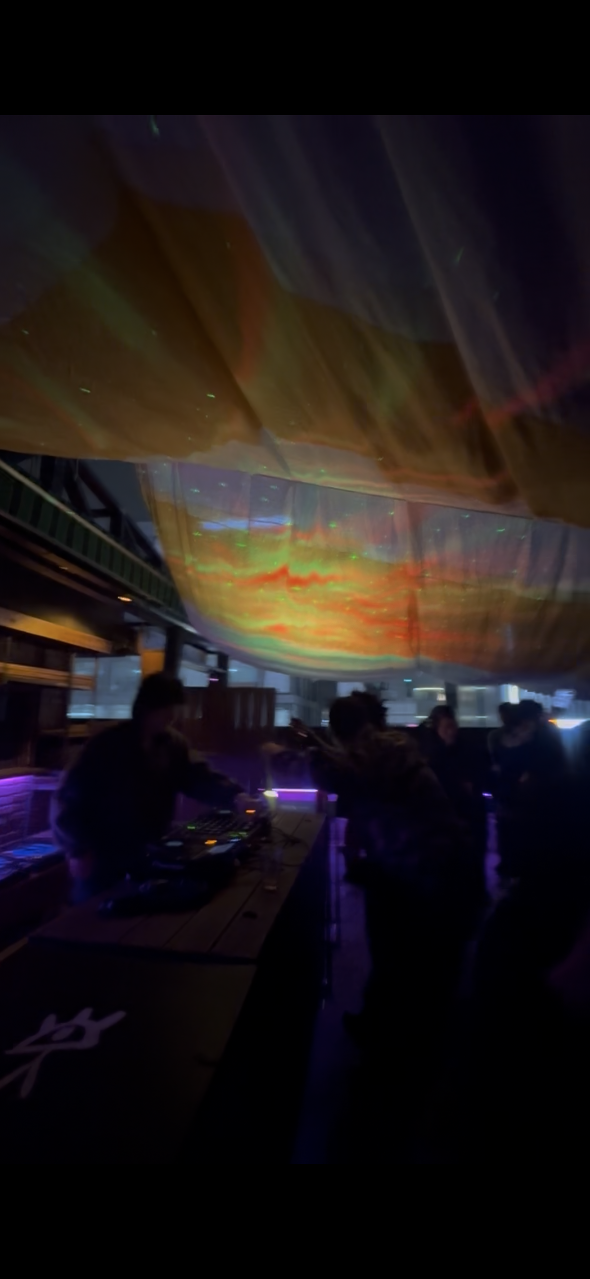 A DJ performs under colorful aurora-like ceiling projections in a dark club.