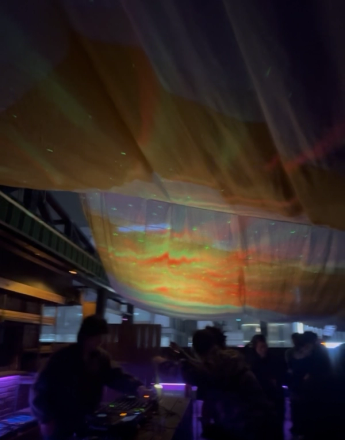 A DJ performs under colorful aurora-like ceiling projections in a dark club.