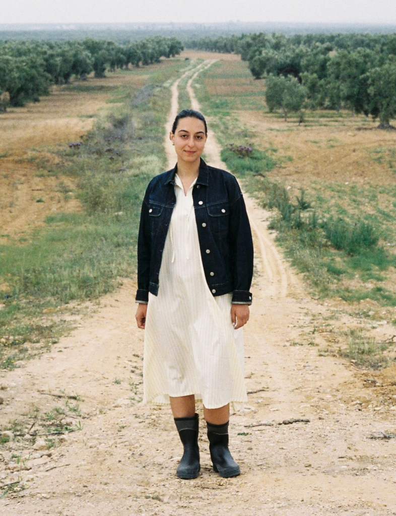 A young woman in a denim jacket, light dress, and rain boots stands on a dirt path in an olive grove.