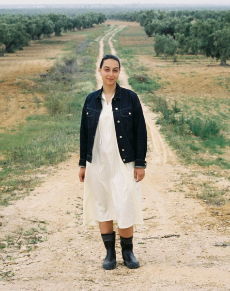 A young woman in a denim jacket, light dress, and rain boots stands on a dirt path in an olive grove.