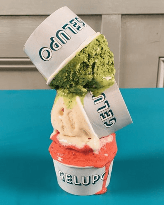 Three stacked and tilted cups of Gelupo gelato in green, cream, and red flavors, melting.