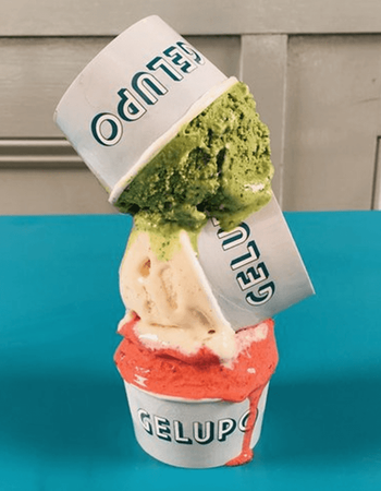 Three stacked and tilted cups of Gelupo gelato in green, cream, and red flavors, melting.