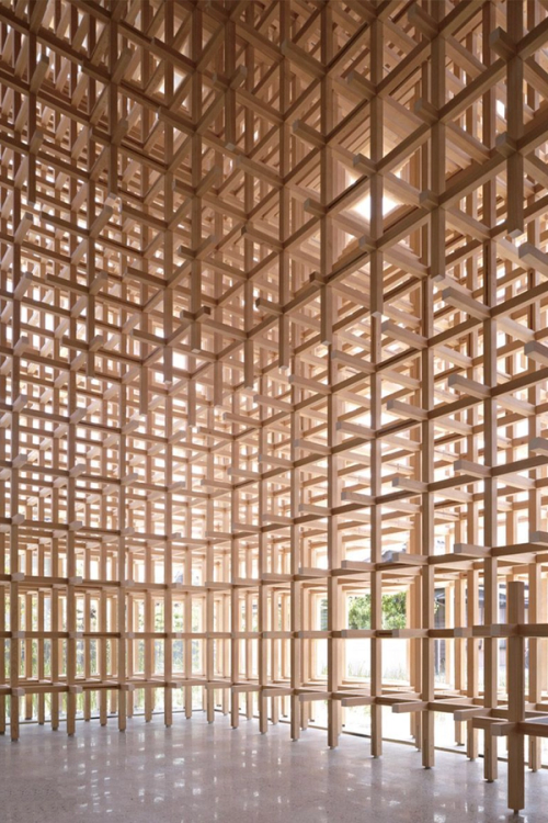 An intricate interior space constructed from a dense, interlocking grid of light wooden beams, with natural light filtering through.