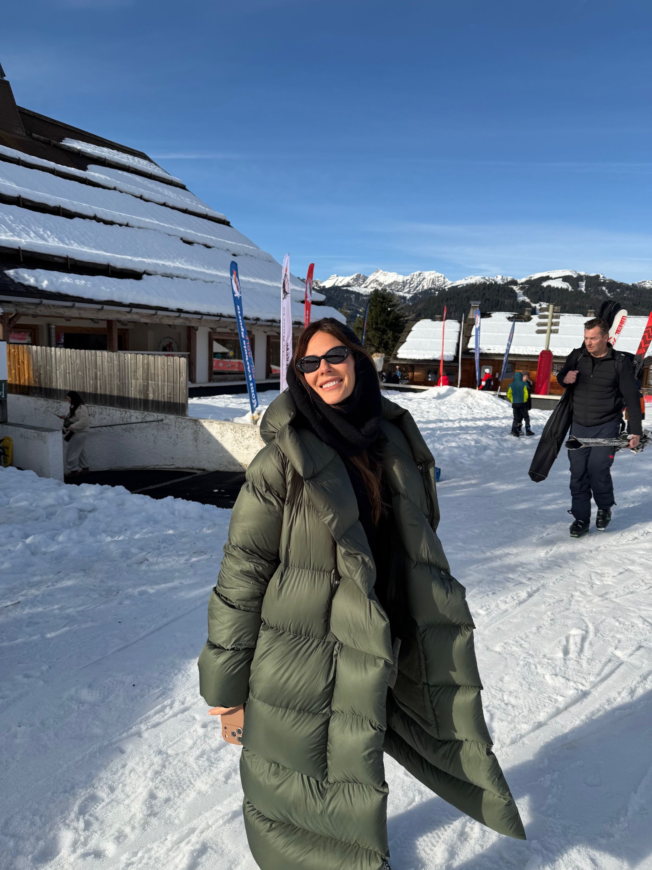 A smiling woman in a long olive green puffer coat and sunglasses stands in the snow at a ski resort.