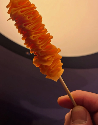 A hand holds a wooden skewer with an elongated, spiraled orange snack.