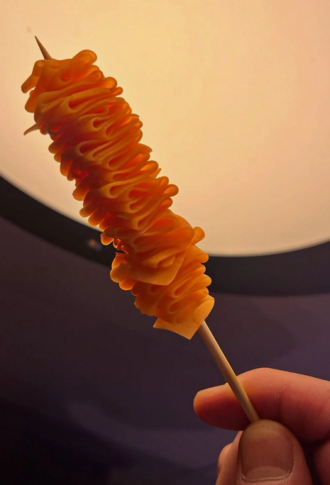 A hand holds a skewer with a deep orange, spiraled snack.