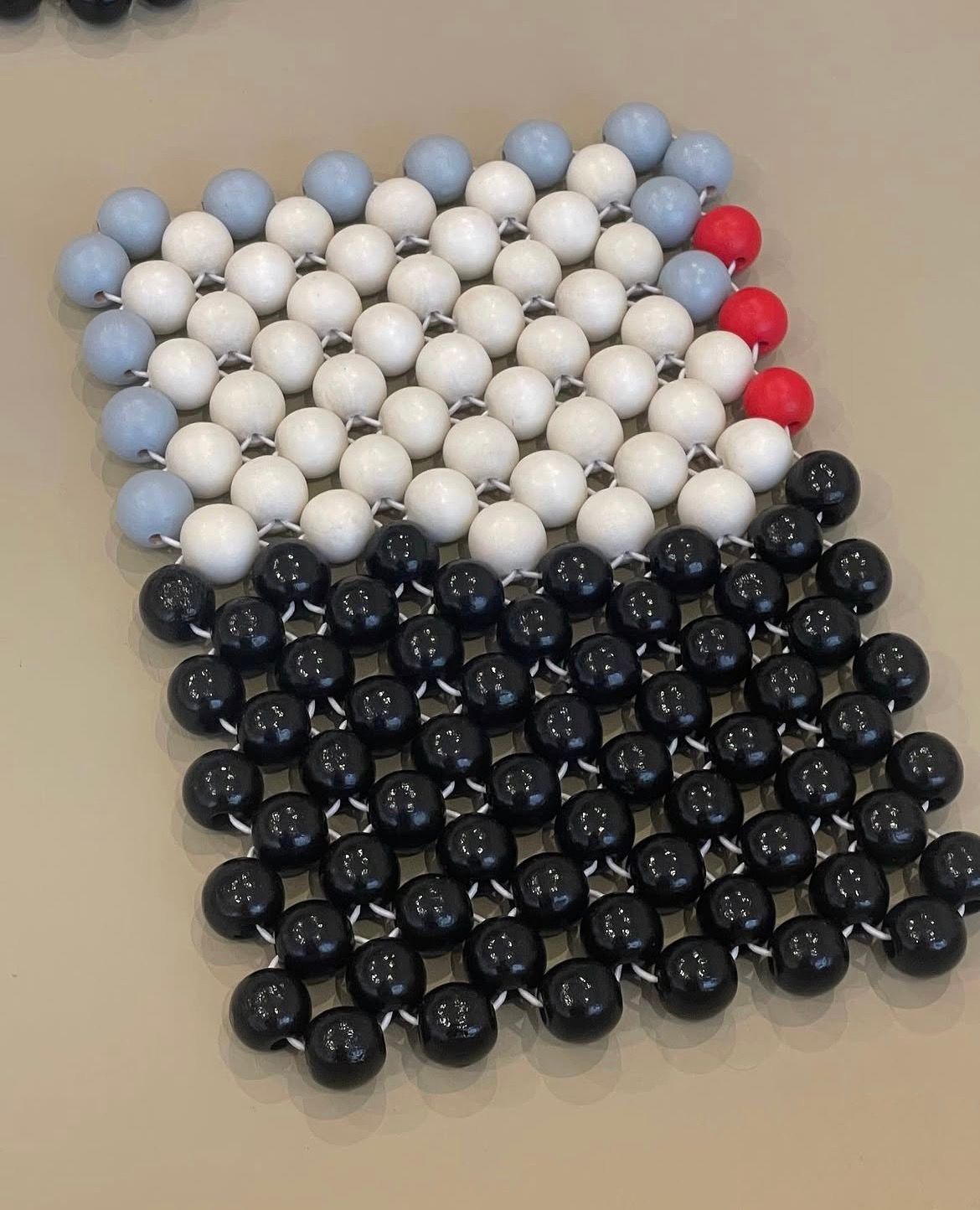 A beaded mat featuring a black bottom half, a wide white band, pale blue beads on the top left, and red beads on the top right.