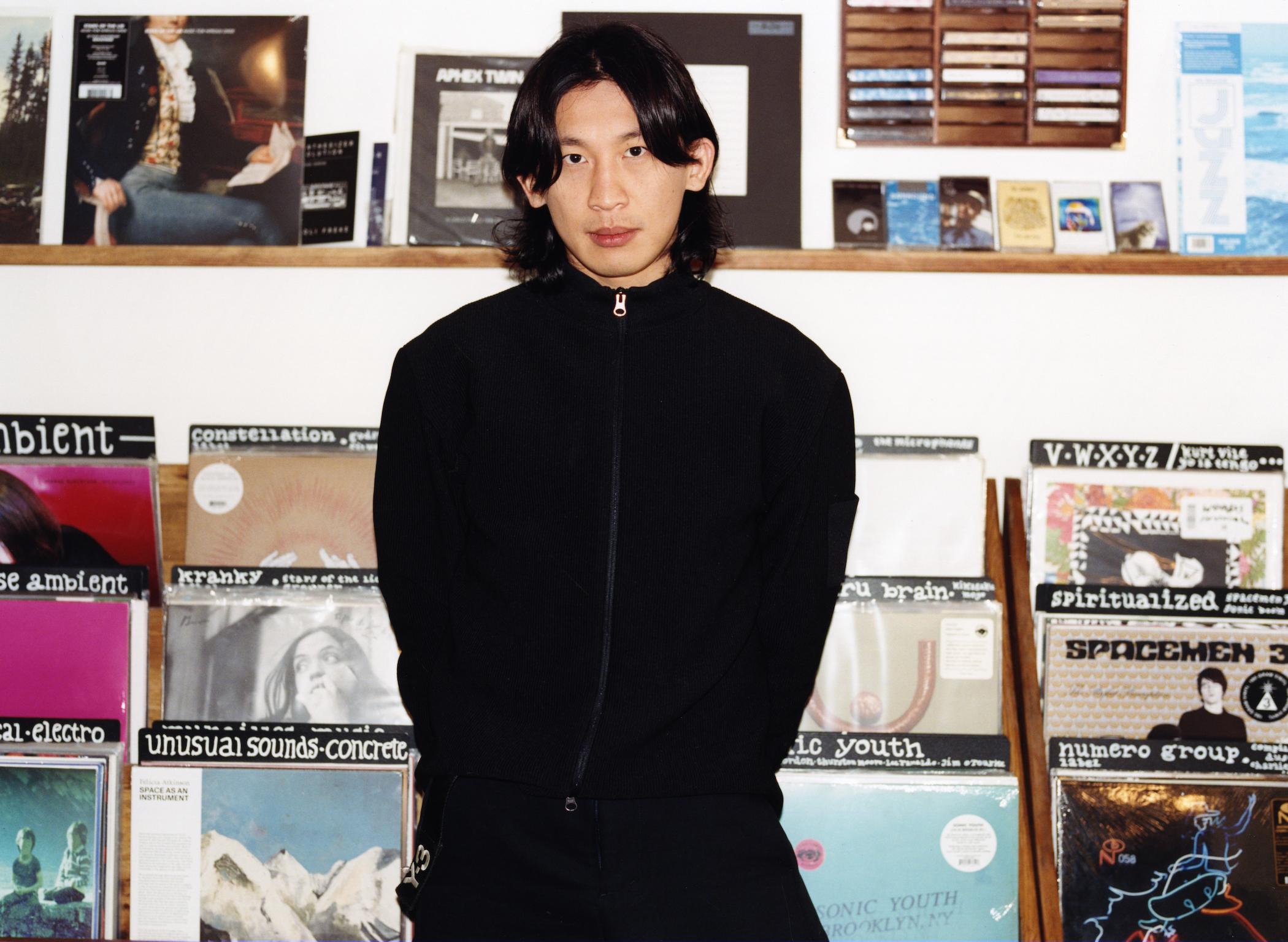 An Asian man with long dark hair stands in a record store in front of shelves filled with vinyl albums.