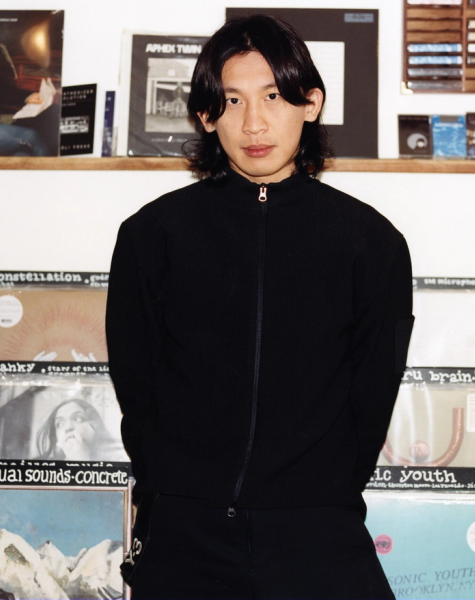 An Asian man with long dark hair stands in a record store in front of shelves filled with vinyl albums.