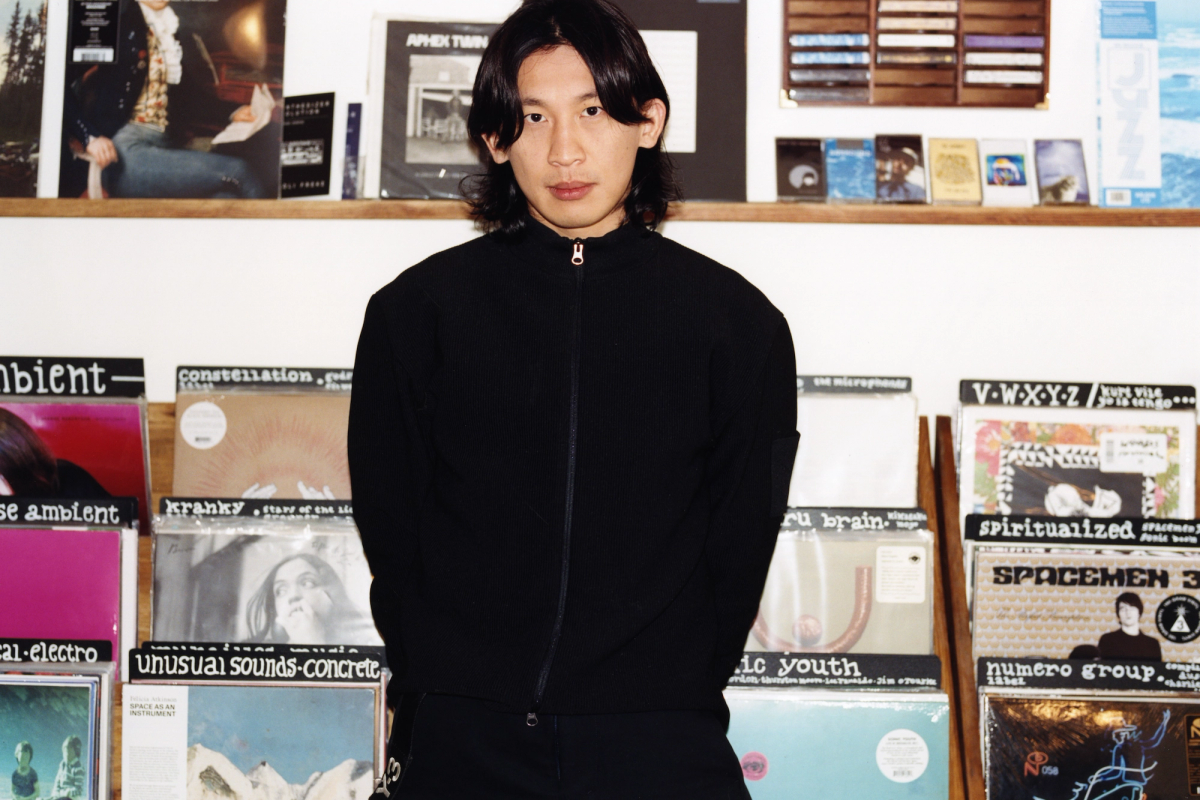 An Asian man with long dark hair stands in a record store in front of shelves filled with vinyl albums.