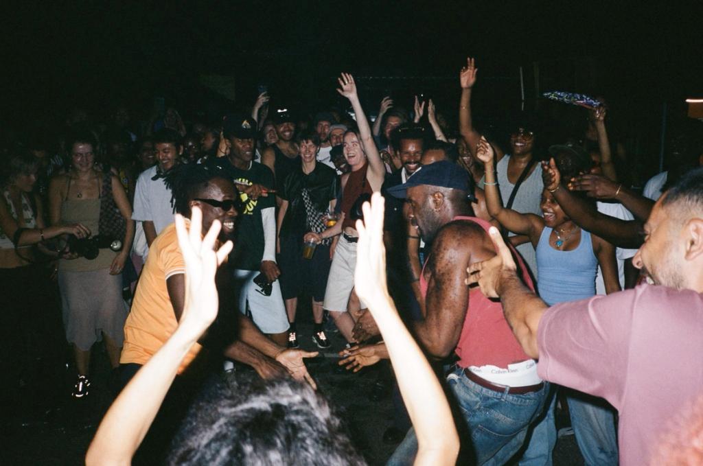 A large group of people dancing energetically in a dark room, with many arms raised.
