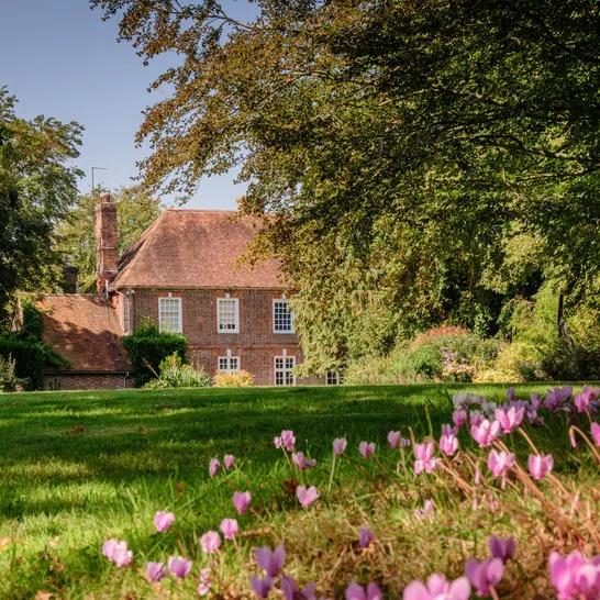 Historic brick house with a red tiled roof, surrounded by green trees and a lawn dotted with pink flowers.