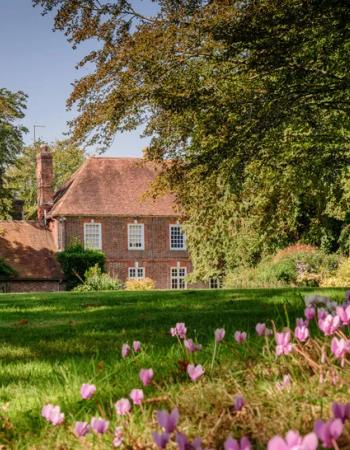 Historic brick house with a red tiled roof, surrounded by green trees and a lawn dotted with pink flowers.