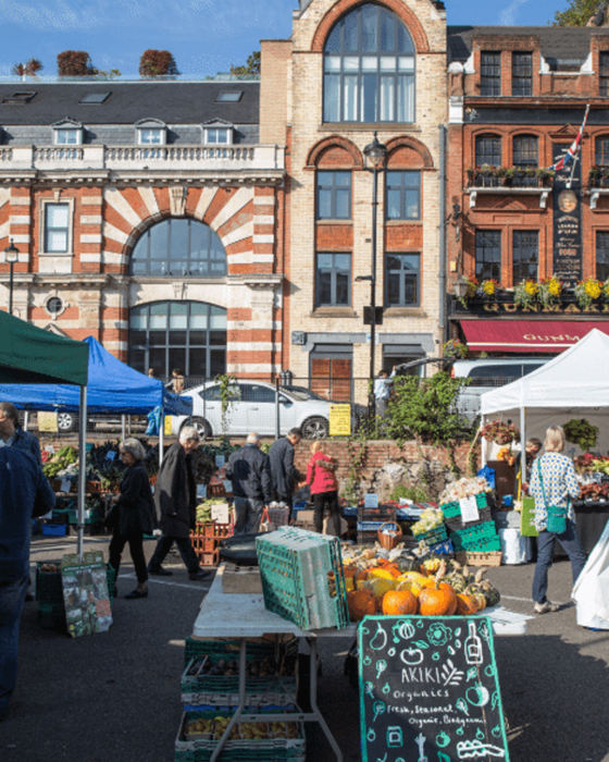 An outdoor market scene with people browsing produce stalls and historic buildings in the background.