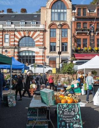 An outdoor market scene with people browsing produce stalls and historic buildings in the background.