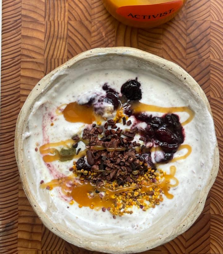 Rustic bowl of yogurt topped with berry compote, honey, cacao nibs, and bee pollen on a wooden surface.