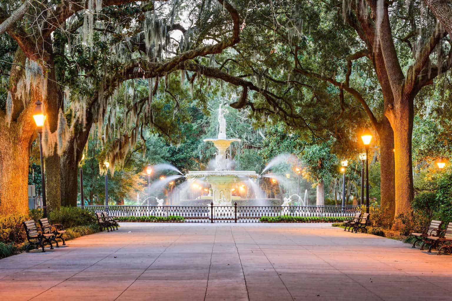 An ornate fountain spraying water in a park at dusk, surrounded by benches, lit lampposts, and trees draped with Spanish moss.