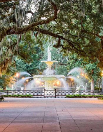 An ornate fountain spraying water in a park at dusk, surrounded by benches, lit lampposts, and trees draped with Spanish moss.