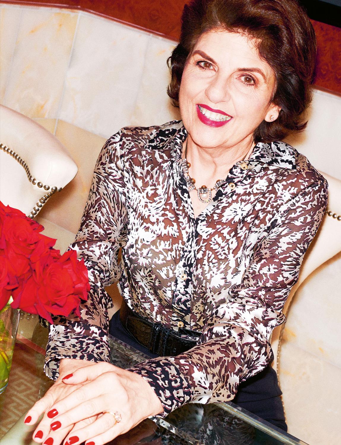 Smiling woman with brown hair, red lipstick, and a patterned blouse, seated.