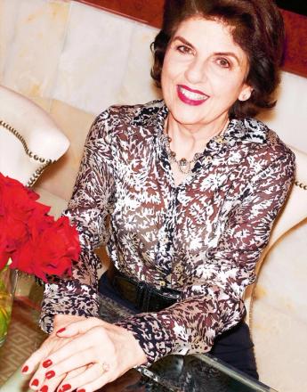 Smiling woman with brown hair, red lipstick, and a patterned blouse, seated.