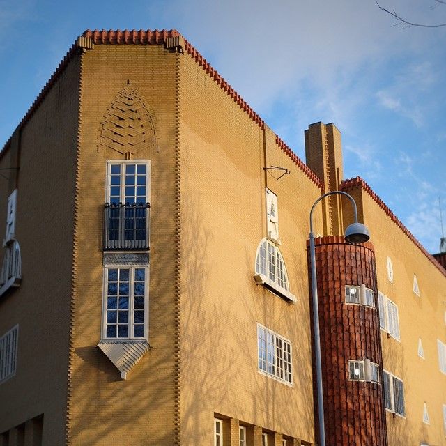 A unique yellow brick building with a red tiled roof, a decorative corner, and a cylindrical tiled structure with an attached street lamp, under sunlight and tree shadows.