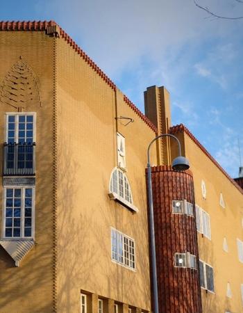 A unique yellow brick building with a red tiled roof, a decorative corner, and a cylindrical tiled structure with an attached street lamp, under sunlight and tree shadows.