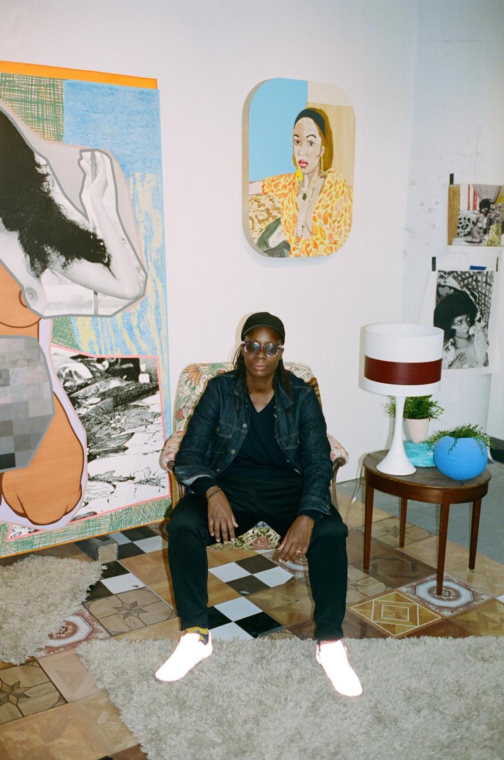 A person in a black cap, clear glasses, and a denim jacket sits in a patterned armchair, facing the viewer. They are in an art-filled room with colorful paintings, including a large abstract work and a portrait, alongside a lamp and plants.