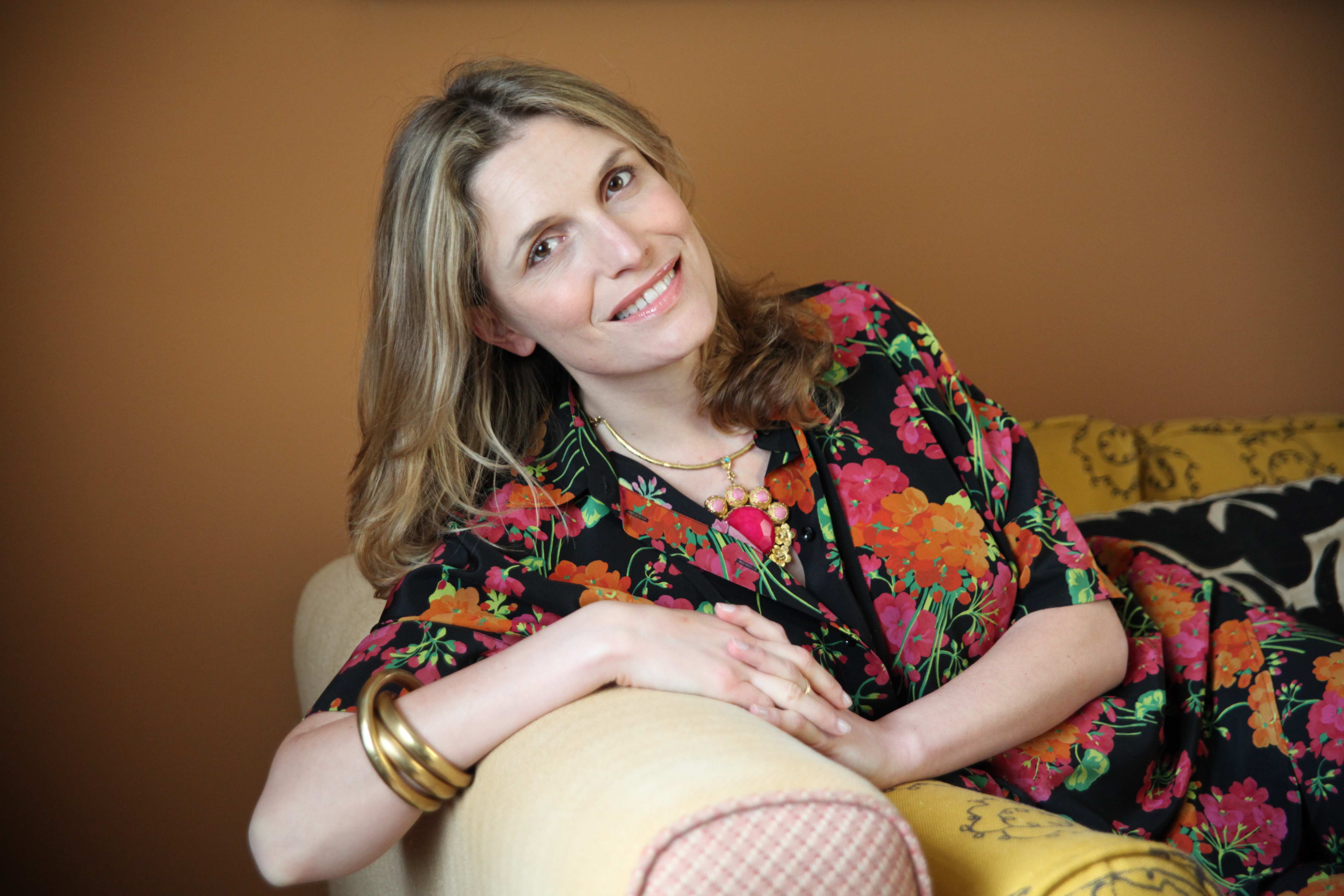 A smiling woman with blonde hair in a black floral dress and gold necklace leans on a couch.