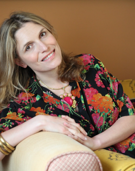 A smiling woman with blonde hair in a black floral dress and gold necklace leans on a couch.
